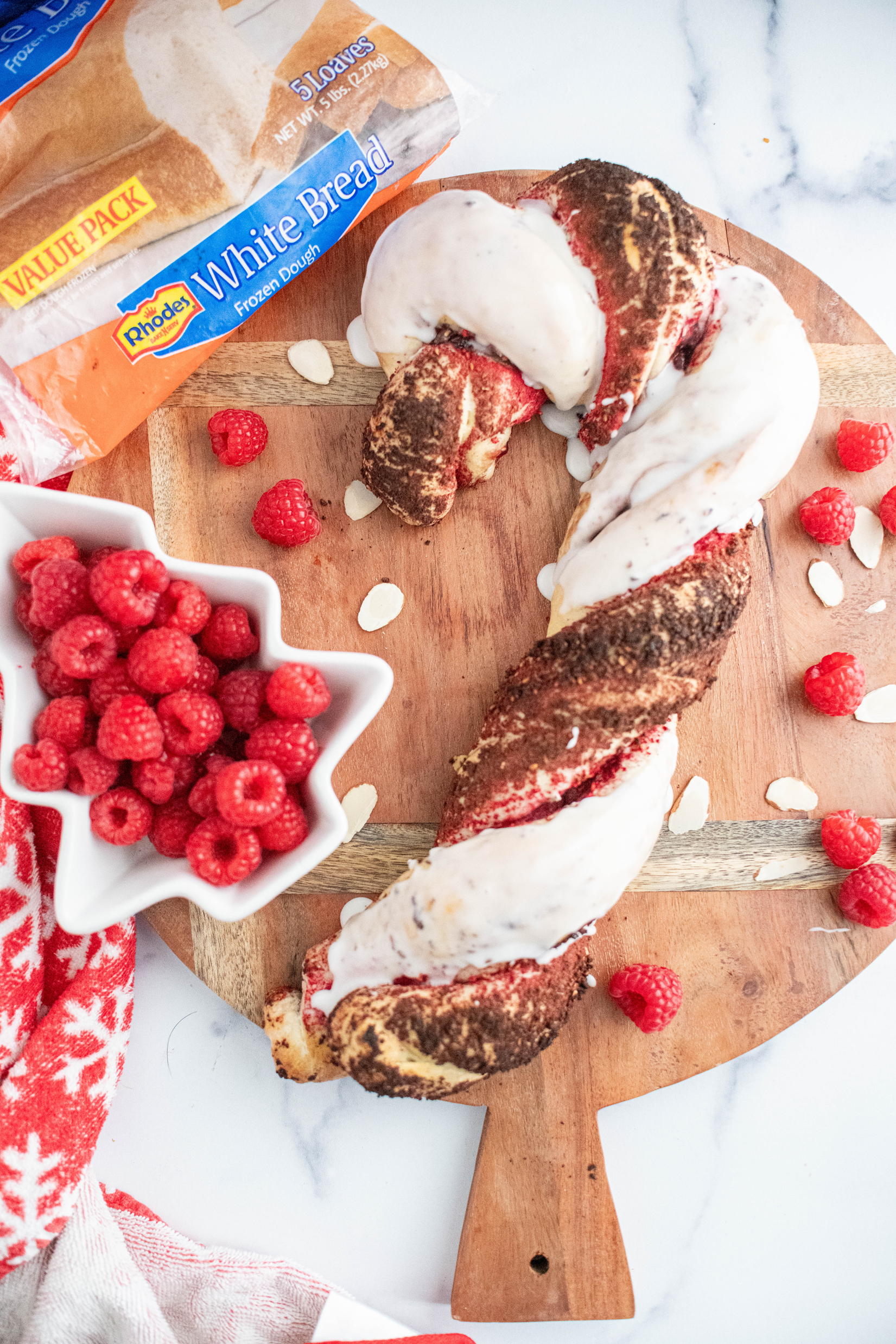 Braided Candy Cane Bread filled with raspberry preserves and sliced almonds, topped with white glaze. Candy Cane bread sitting on a round wooden board.